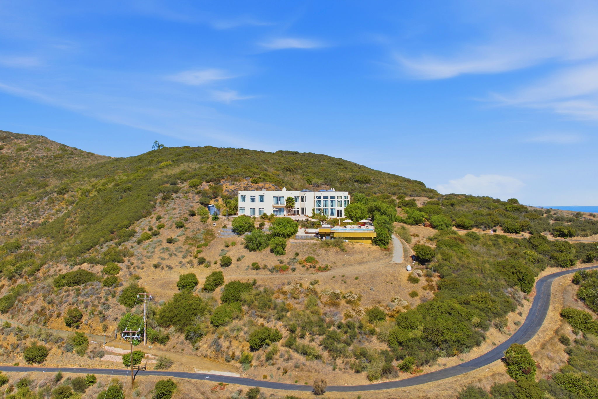Aerial view of the estate alone on its hilltop above the winding road, ringed by protected hillside with the Pacific beyond
